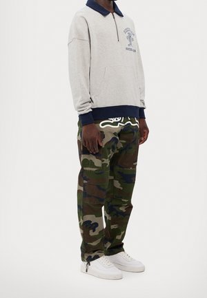 Person trägt einen grauen Pullover mit marineblauem Kragen und camouflage-farbene Cargo-Hose, steht leicht nach rechts gewandt, weiße Turnschuhe sichtbar.