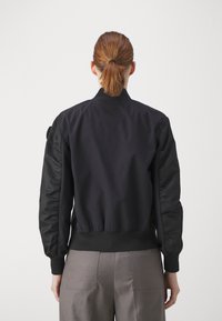 Černá bomber bunda s žebrovaným límcem a manžetami, obsahující látkové panely a hladkou texturu. Záda mají minimalistický design.