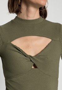 Haut côtelé vert olive avec un col montant, un détail torsadé à l'avant et des sections découpées sous les bras.