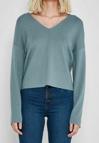 Maglione a collo a V, a maniche lunghe in verde teal smorzato, realizzato in morbido materiale lavorato a maglia. Design cropped con orlo dritto, indossato sopra jeans a vita alta.