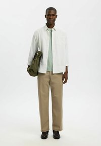 Chemise boutonnée gris clair portée sur un pull vert menthe, associée à un pantalon beige et des chaussures foncées. Tenant un sac vert olive.