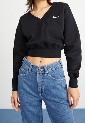 Sudadera negra recortada con cuello en V y logo blanco de Nike swoosh, usada con jeans azules de cintura alta en una persona de pie.