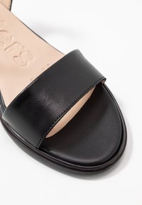 Musta nahkasandaali, jossa on leveä, litteä hihna jalan päällä, sileä pinta, pyöreä kärki ja vaalea pohja, jossa on kohokuvioitu logo.