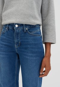 Jeans en denim bleu foncé avec une coupe ajustée, des poches avant et une fermeture à bouton ; associés à un haut à manches longues gris clair.