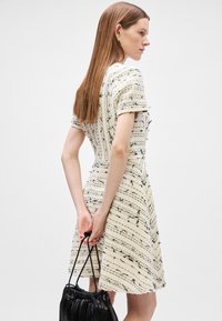 Īsām piedurkņu kleita krēmveida faktūras audumā ar melnām svītrām. Modelis tur melnu plektu maisiņu ar aukliņu, mati krīt taisni līdz pleciem.