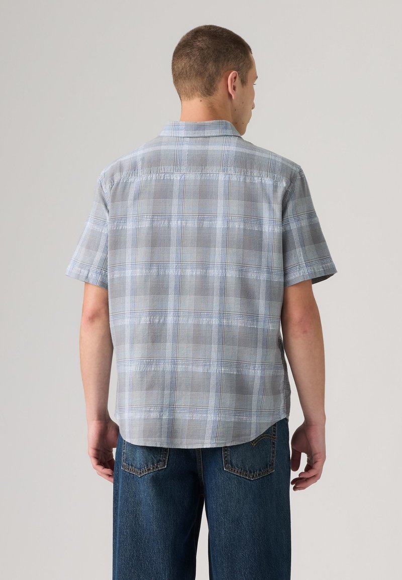 Homme de dos portant une chemise à carreaux gris clair à manches courtes et un jean en denim bleu sur un fond uni.