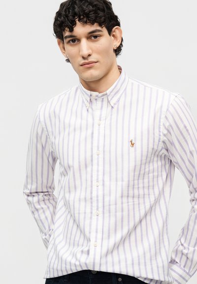 Νεαρός άνδρας που φοράει λευκό πουκάμισο με κουμπιά, με ανοιχτό μωβ κάθετες ρίγες και ένα μικρό κεντημένο λογότυπο στο στήθος.