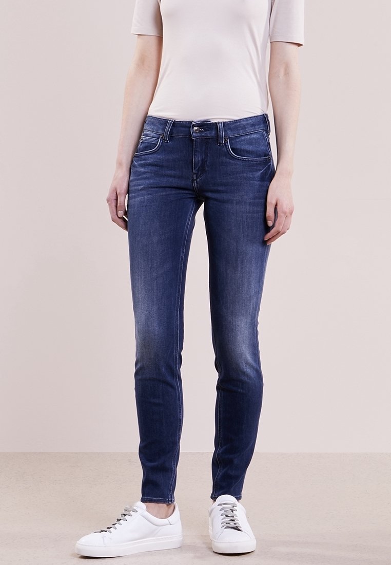 Jeans ajustados de mezclilla en azul oscuro, con cinco bolsillos, ligeras decoloraciones y cierre de botón. Combinados con zapatillas blancas.