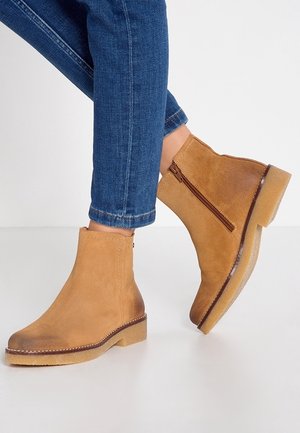 Bottines en daim beige avec une fermeture éclair sur le côté, bout arrondi et semelle en caoutchouc léger et plat. Détails de couture le long du bord.