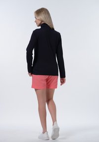 Haut à manches longues bleu marine avec un design texturé, associé à un short corail. Modèle portant des baskets blanches, tourné dos à l'appareil photo.