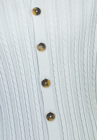 Valkoinen neulecardigan, jossa on johdinmalli, kuusi pyöreää kontrastinväristä nappia ruskeassa ja mustassa sävyssä, sekä teksturoitu ribbidesign.