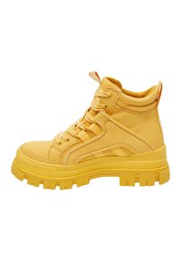 Zapatillas amarillas de caña alta, hechas de lona, con una suela gruesa, cordones en la parte frontal y un tirador en la parte trasera. Tela texturizada con rayas.