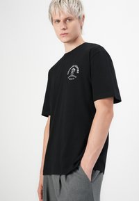 Junger Mann mit kurzen blonden Haaren, der ein schwarzes T-Shirt mit weißem "Cortado Coffee"-Logo und graue Hosen trägt, mit Händen in den Taschen.