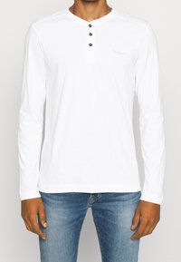 Biała bluza z długim rękawem z dekoltem henley, z trzema czarnymi guzikami i subtelnym detalem logo na piersi. Wykonana z miękkiego materiału.