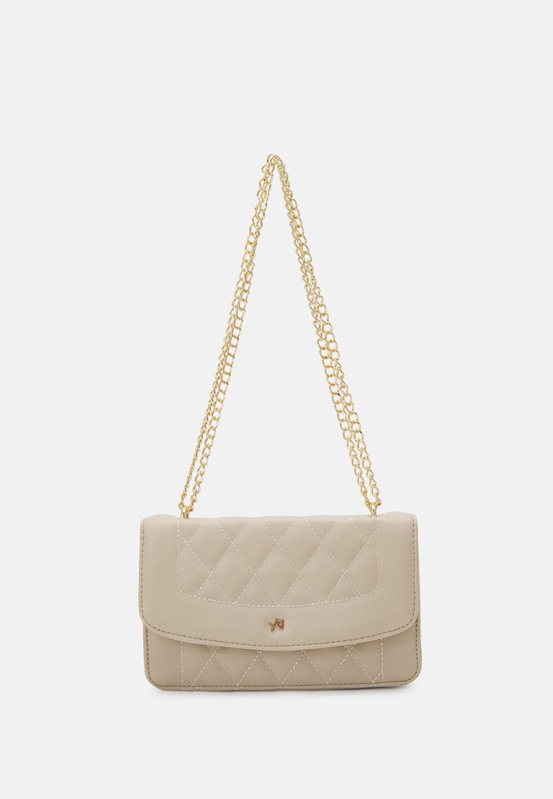 Sac à main matelassé beige avec rabat et petit emblème doré, doté d'une longue bandoulière chaîne dorée pour un porté épaule ou travers.