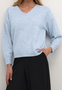 Kobieta ubrana w jasnoniebieski sweter z dekoltem w serek w drobne plamki oraz czarne spodnie, z jedną ręką w kieszeni, a drugą swobodnie opuszczoną wzdłuż ciała.