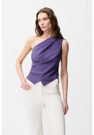 Femme portant un top asymétrique violet sans manches et un pantalon blanc taille haute, debout avec une main sur la hanche.