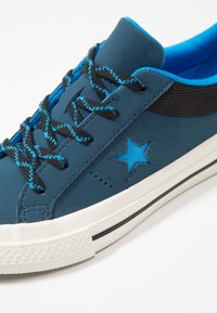 Modrá kožená teniska s černými síťovanými detaily, modrými tkaničkami s vzorovaným designem a bílou gumovou podrážkou s strukturovanou špičkou.