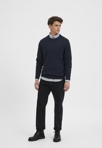 Sudadera de lana en azul marino con cuello redondo, superpuesta a una camisa de rayas en azul claro y blanco. Jeans negros rectos y botas de cuero negras.