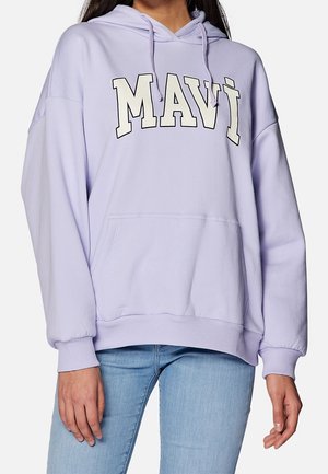 Person iført lys lilla hættetrøje med "MAVI" bogstaver på tværs af brystet og lyseblå jeans, stående med armene afslappet.