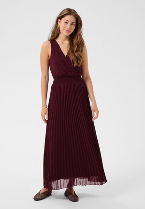 Jeune femme portant une robe longue plissée bordeaux sans manches, des boucles d’oreilles, des bracelets, et des ballerines tissées marron, debout sur un fond blanc.