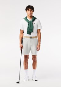 Biele polo tričko s krátkym rukávom, zelený sveter previazaný okolo krku, šedé šortky, béžový opasok, drží čiernu golfovú palicu. Biele ponožky a topánky.