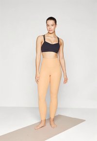 Črna športna modrček z ozkimi naramnico, kombiniran s hlačami breskve barve z visokim pasom. Model stoji na svetlo rjavi joga podlogi pred nevtralnim ozadjem.