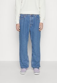 Persoon in losse blauwe spijkerbroeken, een wit langmouwen shirt en paarse en witte geruite instappers, staande tegen een effen achtergrond.