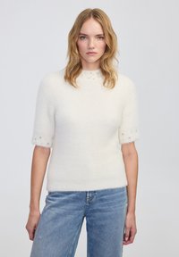 Witte, fluffy trui met korte mouwen en een hoge halslijn, versierd met kralenaccenten op de manchetten. Draagt een blauwe spijkerbroek.