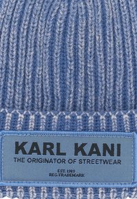 Modrá pletená čiapka s vertikálnou ribovanou textúrou, vybavená modrou látkovou nášivkou s čiernym textom, ktorý znie: "KARL KANI - Pôvodca STREETWEARU."