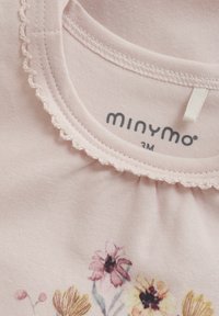 Ljusrosa bomullstopp med vågig halsringning, prydd med blommönster i olika färger och en etikett som visar märket "minymo" och storlek "3M."