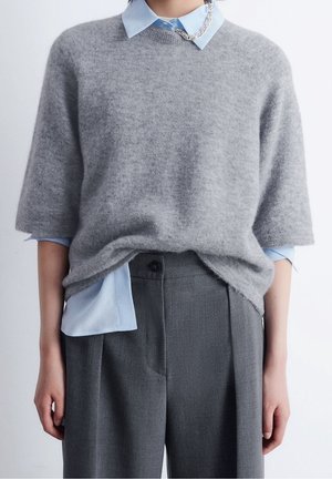 Person trägt ein hellblaues Hemd mit Kragen unter einem grauen, kurzärmeligen, flauschigen Pullover, kombiniert mit hoch taillierten grauen Hosen.