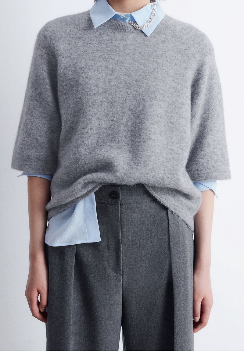 Personne portant une chemise bleu clair à col sous un pull court gris duveteux, associée à un pantalon gris taille haute.