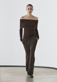 Brown off-the-shoulder top with loose draping and long sleeves paired with tailored, straight-leg brown pants featuring a front pocket detail.