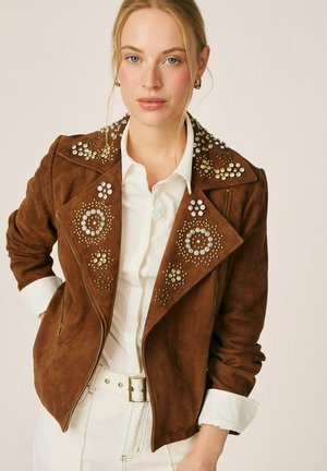 Mujer que lleva una chaqueta de ante marrón con tachuelas redondas decorativas sobre una camisa blanca abotonada y pantalones blancos con cinturón.
