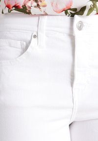 Jeans de mezclilla blanca con un bolsillo frontal, botón plateado y cierre de cremallera, que presentan una textura suave y un ajuste entallado.
