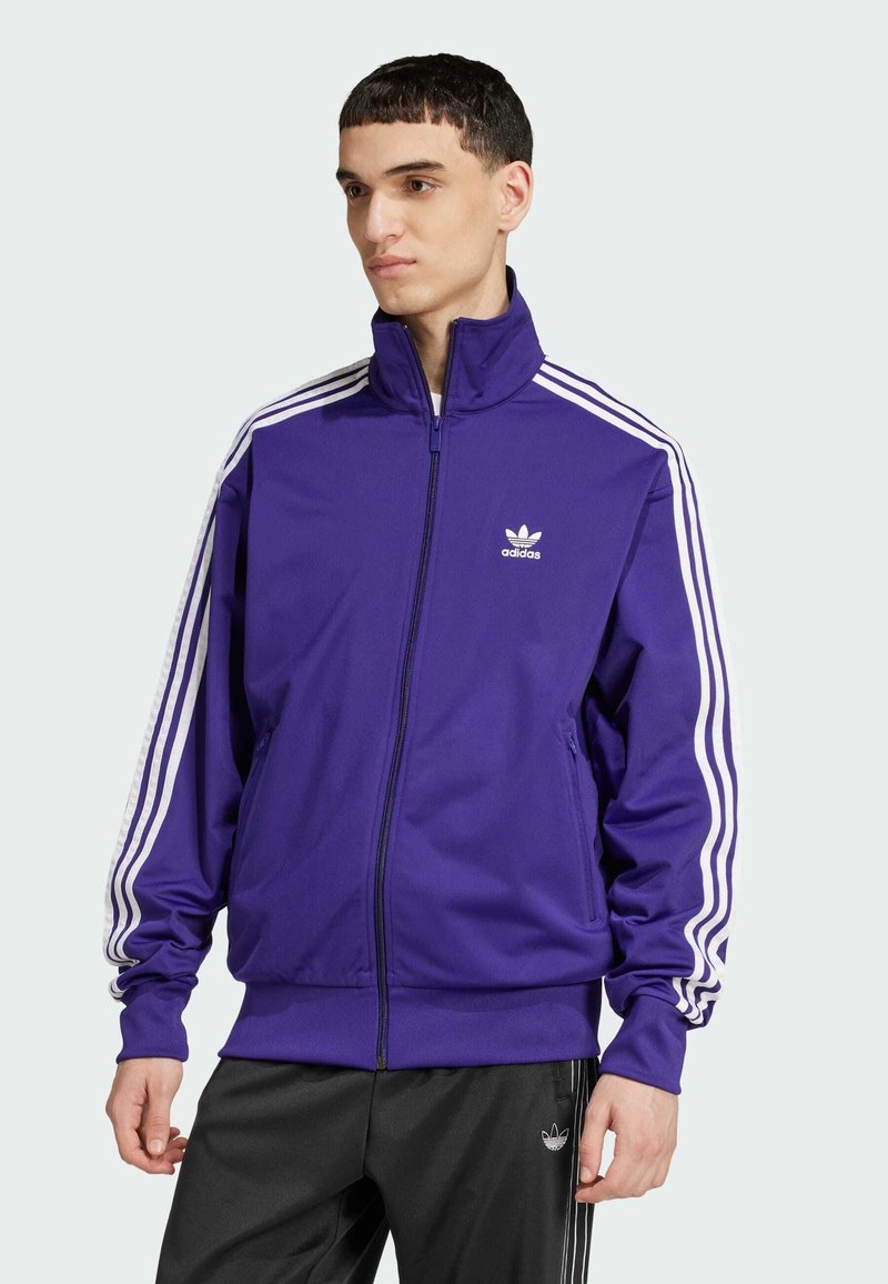 Lilla treningsjakke med høy krage og full glidelås. Har hvite tre-stripe detaljer på ermene og en liten hvit logo på brystet.
