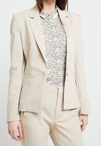 Blazer beige ajusté et pantalon assorti portés sur une blouse blanche avec un motif abstrait de lettres beige et marron.