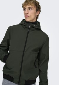 Giacca verde scuro con cappuccio, zip frontale, due tasche laterali e polsini elasticizzati. Texture liscia con design moderno e aderente.