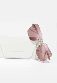 Óculos de sol Michael Kors com lentes rosa angulares e detalhes em dourado, repousando sobre uma caixa branca texturizada, apresentando o nome da marca em relevo na parte frontal.