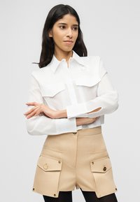 Γυναίκα με μακριά σκούρα μαλλιά φορά διαφανές λευκό πουκάμισο με κουμπιά και μπεζ σορτς ψηλόμεσο με μεγάλες τσέπες με καπάκι, φόντο γκρι.