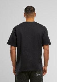 Camiseta negra de algodón con cuello redondo y mangas cortas. Presenta un diseño sencillo y se combina con pantalones oscuros que muestran un detalle de logo.
