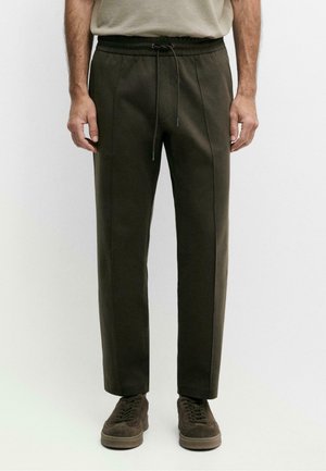 Uomo che indossa pantaloni verde oliva scuro con coulisse abbinati a sneakers in suede marrone, in piedi davanti a uno sfondo semplice.