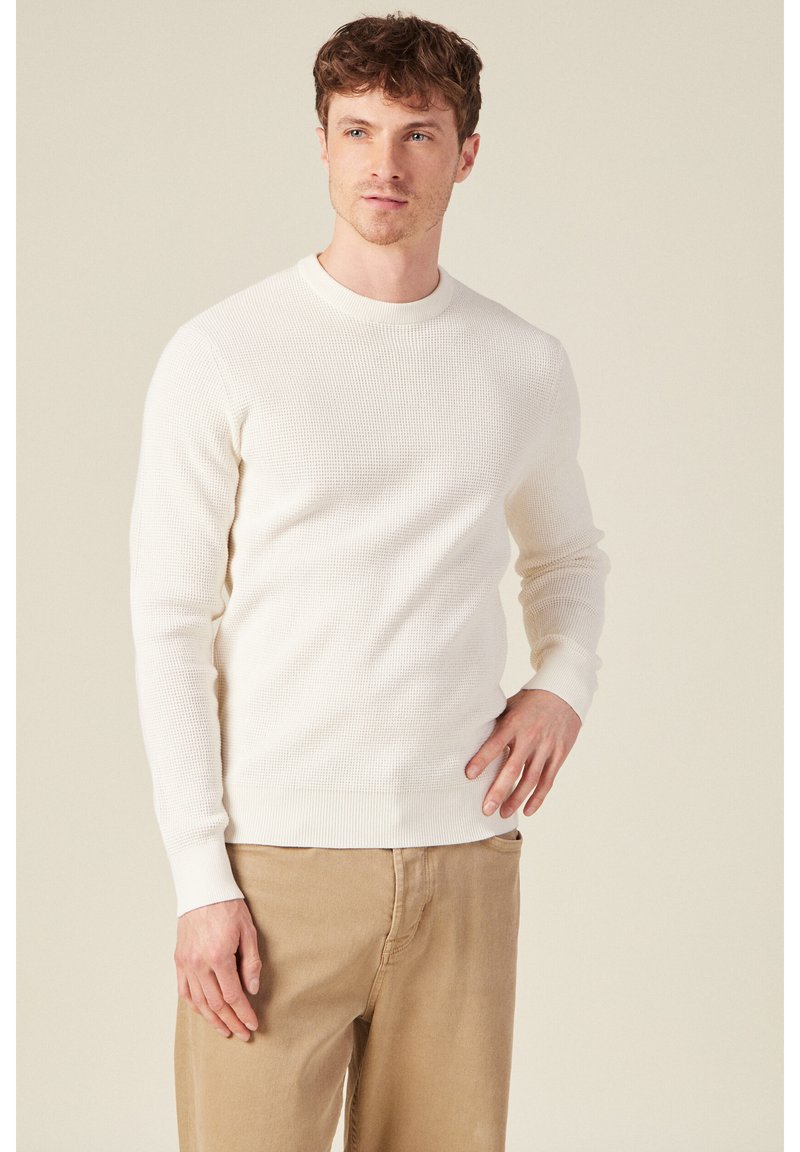 Weißer Strickpullover mit Rundhalsausschnitt und gerippter Textur, kombiniert mit hellbeigen Hosen. Der Schnitt ist schmal, mit langen Ärmeln und gerippten Bündchen.