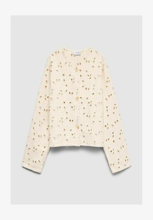 Blouse crème à manches longues boutonnée avec broderie florale ajourée et col rond sur fond uni clair.