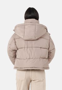 Beige pufferjacka med huva, med ett quiltat mönster och synliga sömmar. Slät yta med en avslappnad silhuett och kort längd.