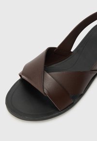 Braune Leder-Sandale mit einem Design aus Kreuzriemen, strukturierter Oberfläche und einer mattschwarzen Gummisohle. Verfügt über eine offene Ferse für einfaches Anziehen.