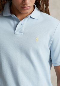 Ljusblå pikétröja med texturerat tyg, som har en treknappsknäppning och en liten, gul logotyp på vänster bröst.