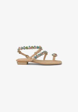 Sandalias planas beige con tiras joyadas que presentan piedras multicolores. El diseño incluye un pequeño lazo para el dedo del pie y un tacón cuadrado con acentos dorados.