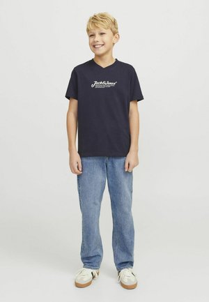 T-shirt en coton bleu marine avec un décolleté en V et un logo blanc, associé à un jean en denim bleu clair et des baskets crème avec des accents verts.